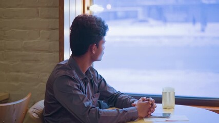 Young man sitting alone by window in cafe, staring outside at snowy street, hands clasped on table, wearing formal shirt, looking contemplative, natural light illuminating indoor winter setting