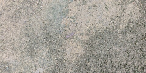 Rough soil with small stones, fine sand, and scattered grass. The surface is uneven, showing natural texture with brown, gray, and light green tones.