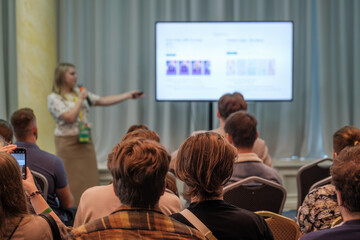 Presenter speaks into microphone while pointing to projected slide as attendees watch. Bright room, conference setting, engaged audience, learning vibe, professional presentation, sharing information.