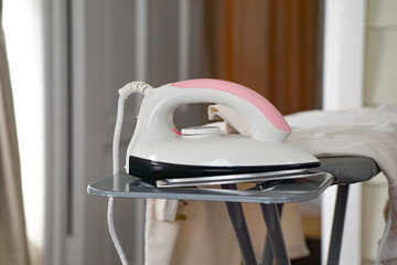 A white and pink electric iron rests on the ironing board, with beige curtains and a closed door in the background.