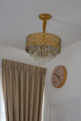 A stylish room corner featuring a gold chandelier with crystal pendants, pleated beige curtains, and a round wall clock with a pink face and gold frame.