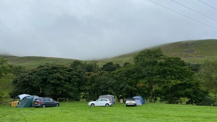 Scenic countryside camping area with tents and cars on a green field surrounded by trees and misty hills in the background. Peaceful outdoor travel and adventure concept in rural England.