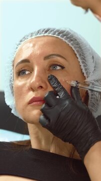 Middle years woman receiving cosmetic injection in cheek area for skin rejuvenation, vertical orientation. Female patient undergoing anti-aging treatment with dermal filler at aesthetic clinic. Mature