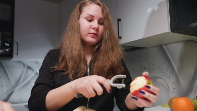 Young lady in modern kitchen peels red apple with handheld peeler, focused expression, blue nails visible, fruit prep under bright light, fresh snack routine, clean countertop background