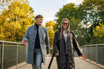 Smiling man and woman walk side by side on a bridge holding fall leaves, wearing long coats and sunglasses, surrounded by green and golden trees. No visible logos