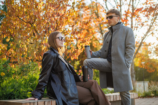 Stylish man and woman in coats and sunglasses enjoy warm drinks on a park bench surrounded by golden fall trees