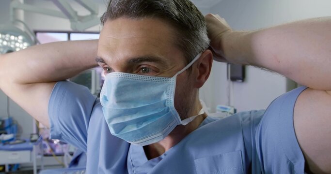 Pulling mask straps scrub-clad surgeon preparing in operating room, under surgical light