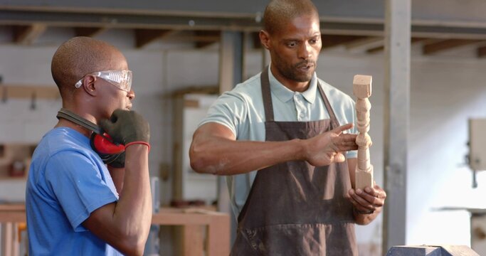 Showing mentor guiding apprentice examining wooden spindle at woodshop bench, with safety goggles