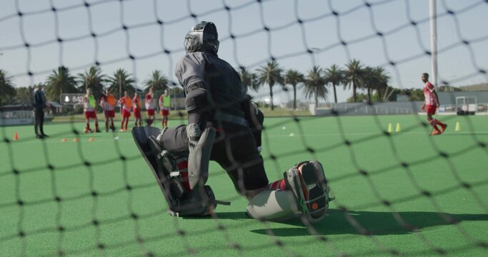 Kneeling field hockey goalkeeper wearing helmet and leg guards defending net on turf, with cones
