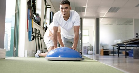 Kneeling man wearing white shirt and shorts doing balance drill on green turf, with blue BOSU