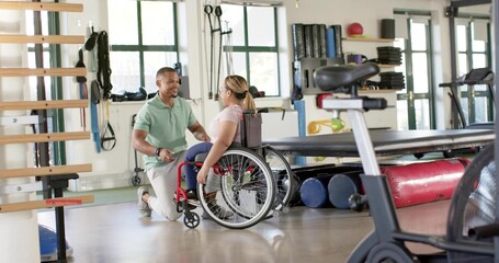 Kneeling therapist in green polo helping manual wheelchair client in rehab gym by bike, copy space