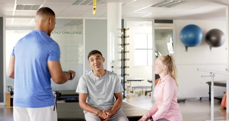 Standing trainer in blue shirt holding timer and instructing seated clients on treatment table, TRX
