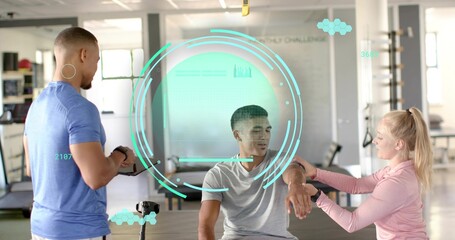 Seated client receiving forearm check in gym studio, trainers in athletic shirts using tablet HUD
