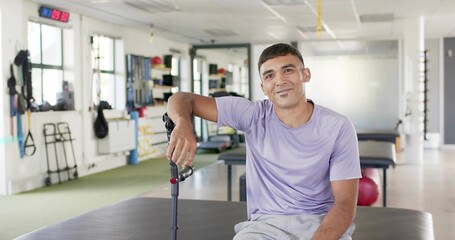 Sitting man in lavender shirt, gray shorts holding black forearm crutch on padded table, clinic