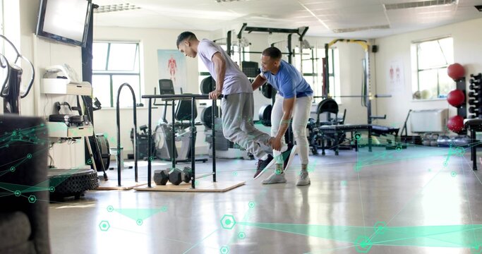 Performing client in light tee gray sweats lowering on parallel dip bars, trainer supporting ankle