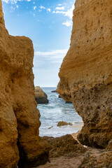 Two sandstone rocks stand on the beach framing a narrow glimpse of the sea beneath a blue sky brushed with wispy clouds