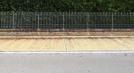 Elegant fence made of metal railing with spikes on brickwall base. High hedge on behind, yellow sidewalk and street in front. Background for copy space.