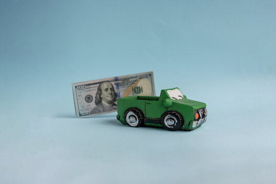 A green toy car is positioned in front of a one hundred dollar bill. The background is a light blue color, creating a playful and financial theme.