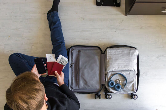 Traveler preparing for trip, holding passport, boarding pass, cash and smartphone near open suitcase, travel planning concept. - Powered by Adobe