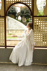 Fototapeta premium Full length female figure of blonde model wearing white fantasy wedding gown. whimsical sitting pose looking out of a window of historical castle location 