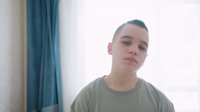 Teen boy practices neck flexibility routine in bright home setting near window with soft daylight, standing still in relaxed posture, he tilts head gently to stretch neck muscles for mobility