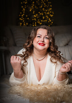A portrait of a beautiful,chubby  woman with a sweet smile against a New Year's backdrop. A buxom woman in a white knitted sweater with an open neckline poses against a backdrop of Christmas lights.