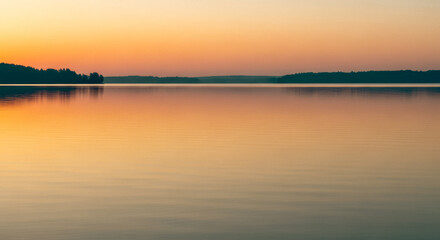 Calm lake at sunset with orange sky reflection. Serene minimalist nature landscape background. Peaceful water and forest silhouette on horizon. Beautiful twilight scene for meditation