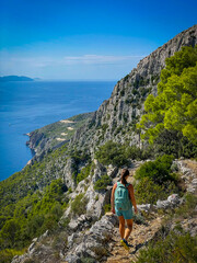 Fit female hiker with backpack walks along narrow cliffside trail with stunning view of blue sea...