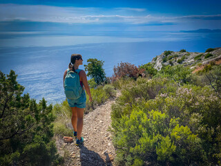 Fit woman and her mixed breed dog hike a sunny coastal path lined with green shrubs and sea views. Summer adventure, blending fitness, nature, and companionship in a peaceful Mediterranean setting.
