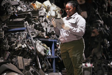 Black female technician check used car damaged engine at scrapyard warehouse recycle area part. African American engineer inspecting rusty oily auto motor old spare part in junkyard for reuse service