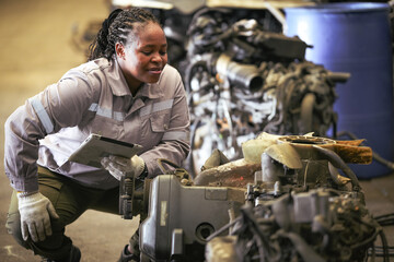 Black female technician check used car damaged engine at scrapyard warehouse recycle area part. African American engineer inspecting rusty oily auto motor old spare part in junkyard for reuse service