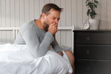 Good Morning. Caucasian Handsome Bearded Guy Sitting On Bed Yawning Covering Face, Mouth with Hand. Young Adult Man Waking Up at Home. Clear White Colors. Sleepless Male Feeling Tired. Sleepy Bachelor