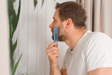 Handsome man combing beard and moustache sitting at home light white bedroom. Guy styling grooming his beard with a comb. Self-care. Softly lit indoor space. Portrait. Bath morning routine. Side view