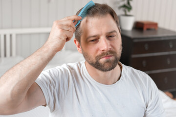 Handsome man combing brushing hair sitting at home light white bedroom. Guy styling grooming his hair with a comb. Self-care. Softly lit indoor space. Portrait. Male haircare morning routine concept
