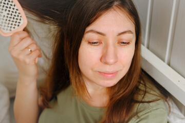 40 years woman takes care combing hair at home in the morning. She brushing hair with a comb. Brunette Female Brushing Hair With Hairbrush Sitting in Bed. Haircare. Candid authentic shot. Real people