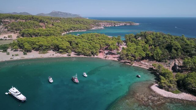 Drone view of Phaselis bay in Antalya, Turkey. Boats anchored in turquoise water near the ancient ruins surrounded by pine forest and mountains.