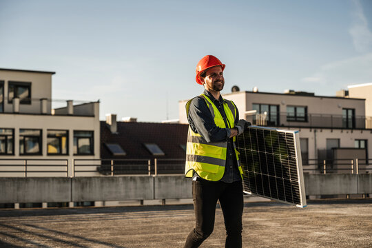 Engineer wearing protective workwear carrying solar panel at terrace on sunny day