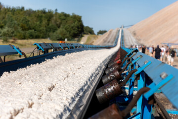 Long blue conveyor belt transports white potash ore material toward massive potassium mining dump sunlight. Industrial machinery stretches distance mineral extraction continuous production process
