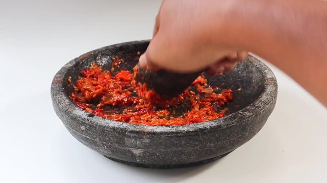 Asian man hands are grinding chilies into traditional Indonesian chili sauce (sambal), using a mortar (cobek) and pestle made of stone. isolated on white background.