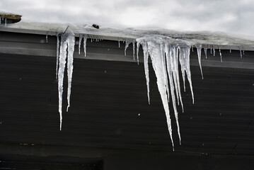 Long frozen icicles along roof eaves creating slippery surfaces and dangerous falling ice hazard for people passing underneath. Sharp icicles dangling from building roof edge, pedestrian safety risk
