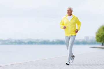 Senior man jogging along a waterfront promenade in a bright yellow hoodie and gray pants, enjoying a healthy lifestyle and the beauty of nature during morning exercise