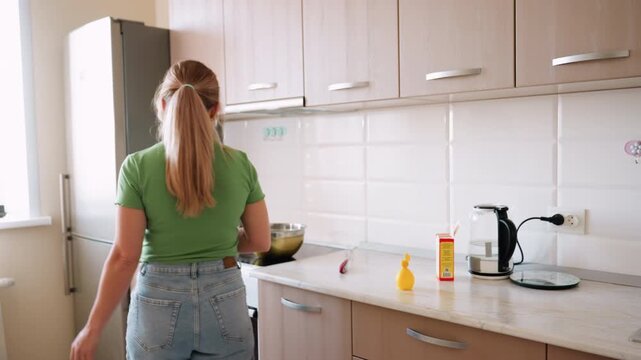 Back view of housewife walking across kitchen toward bowl on burner with decorative light blinking in curtain, creating warm domestic scene of daily cooking and household routine