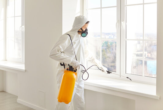 Man wearing protective white ppe suit and face mask disinfecting office workspace or empty apartment and doing pest control with disinfectant spray. Disinfection and protection concept.