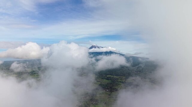 Aerial view of a majestic merapi mountain peak shrouded in clouds, surrounded by lush green landscapes and a serene sky, Klaten Regency, Central Java, Indonesia.