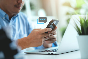 A man is using his cell phone while sitting at a desk with a laptop