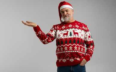 Merry mature man wear knitted christmas sweater Santa hat presenting something on grey. New year shopping idea concept. New Year 2026 Christmas