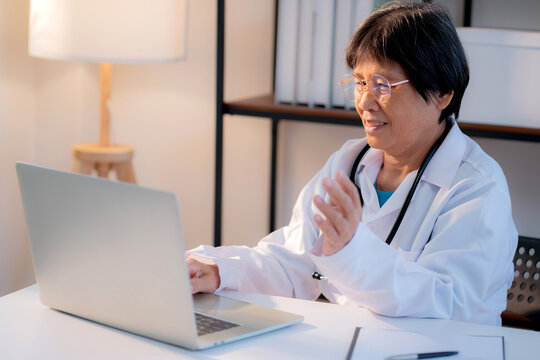 Senior asian doctor woman working on laptop computer on desk in clinic, elderly female physician video call or online consultation with medical, assistance or support with medical.