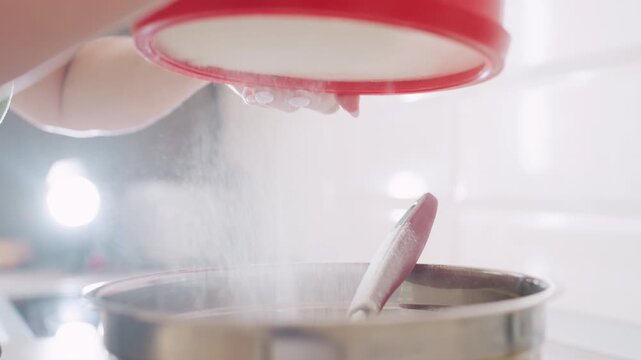 Close up of caterer filtering flour with red sifter into stainless bowl, fine flour dust falling through sieve while mixing utensil rests inside bowl during careful baking preparation in kitchen