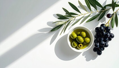 Overhead view of green olives in oil next to dark grapes and olive branch on white surface with bright sunlight casting shadows