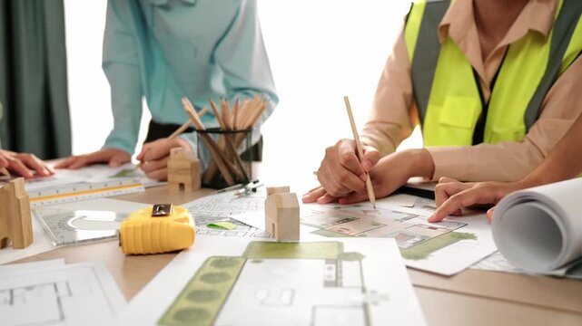 Smart architect and safety engineer discussion about building blueprint on table with architectural document, measurement tools and wooden block scatter around at modern office. Closeup. Delineation.